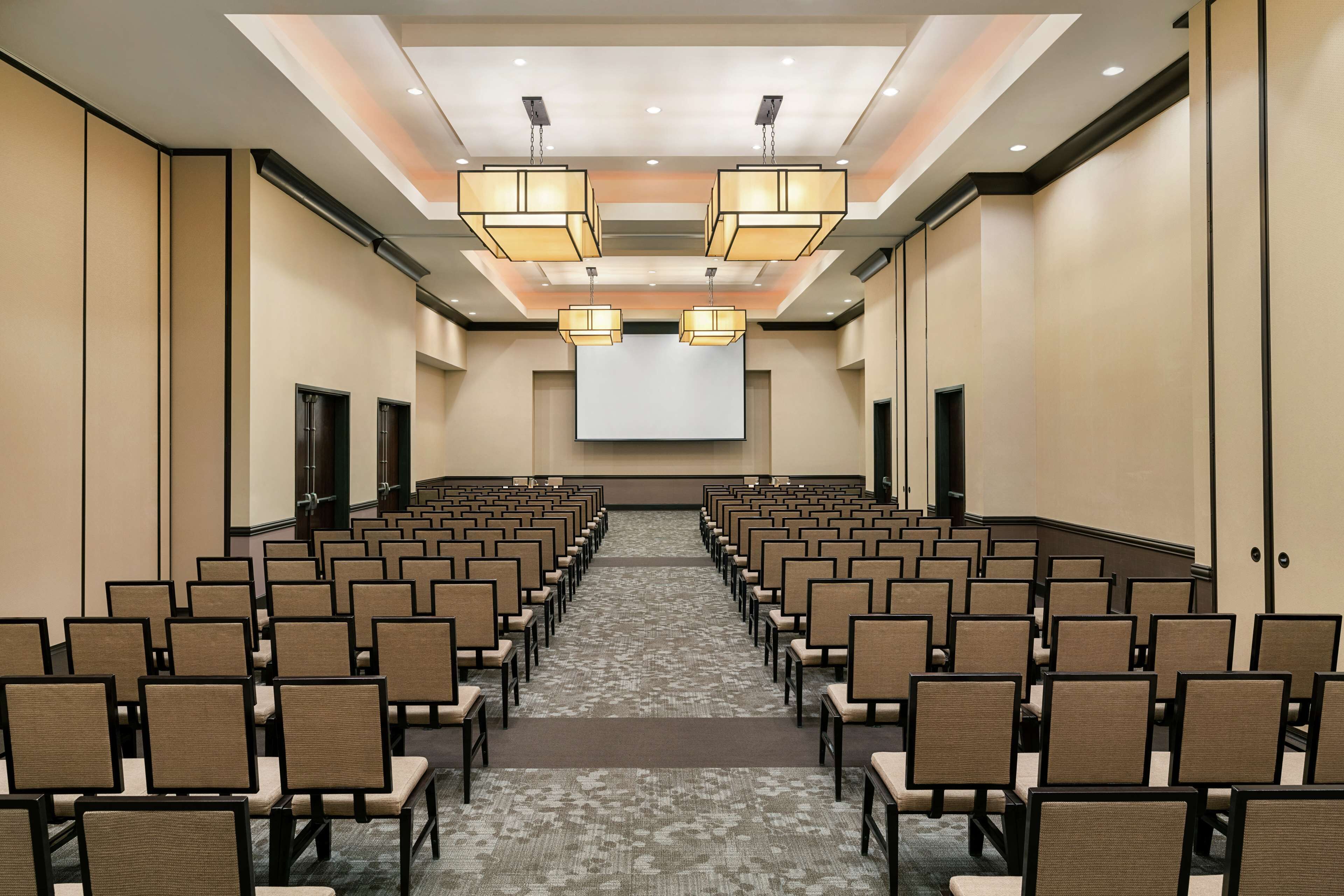 Un salón de conferencias con filas de sillas, una pantalla grande al frente y luces en el techo.