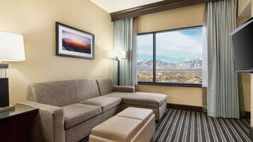Habitación de hotel con sofá, reposapiés y una ventana grande con vistas a la montaña.
