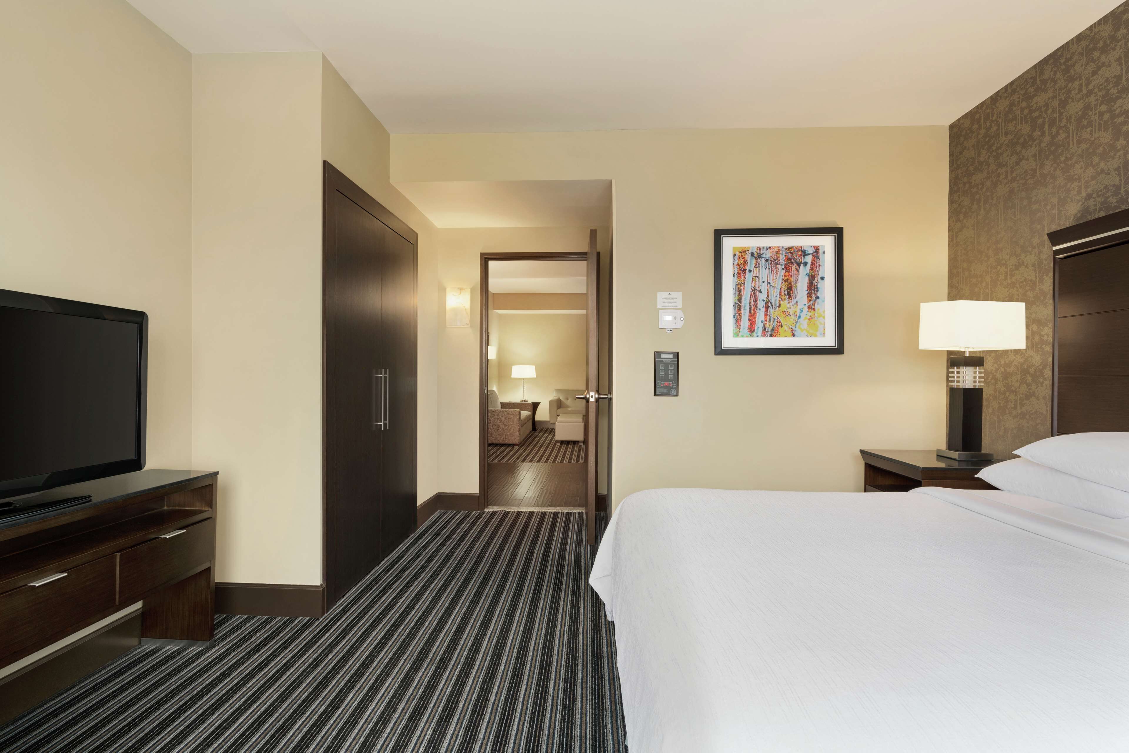 A hotel room with a large bed, a TV on a wooden dresser, a desk with a lamp, and a framed artwork on the wall.