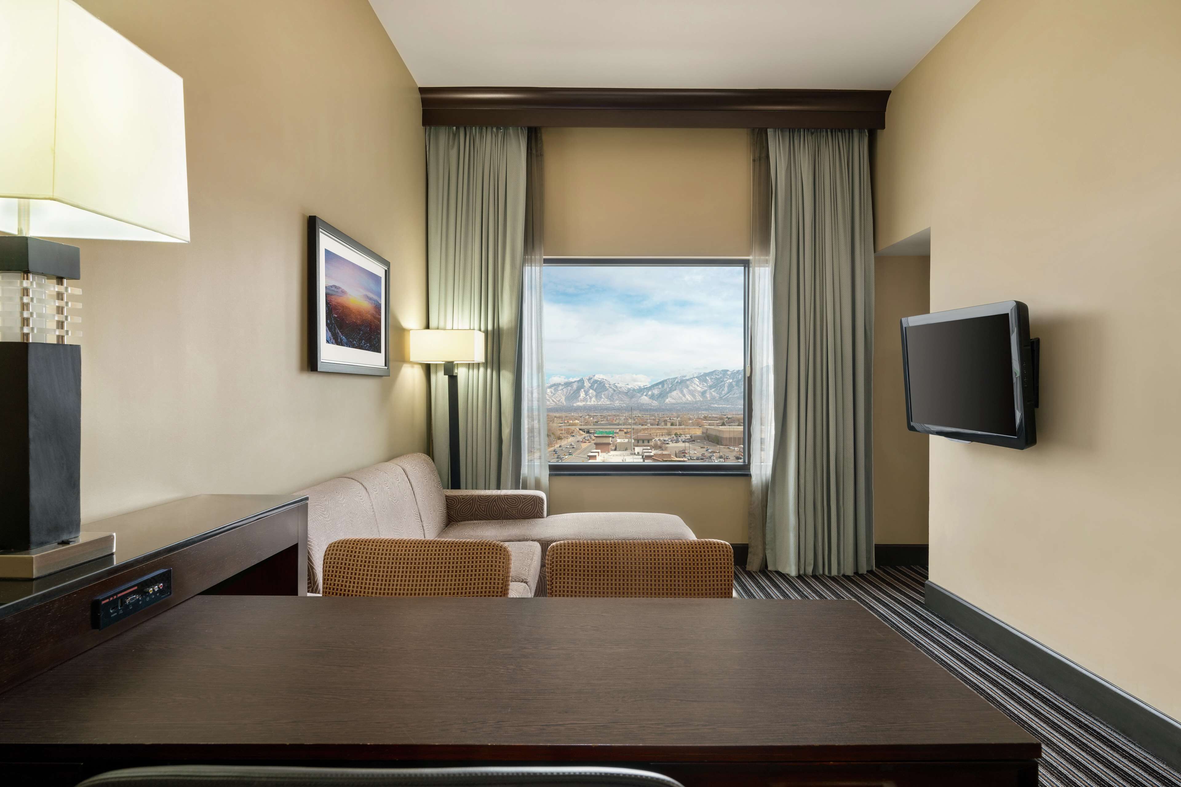 Une chambre d’hôtel dotée d’une grande fenêtre, d’un téléviseur à écran plat, d’un bureau avec une lampe, d’un canapé et de deux fauteuils.