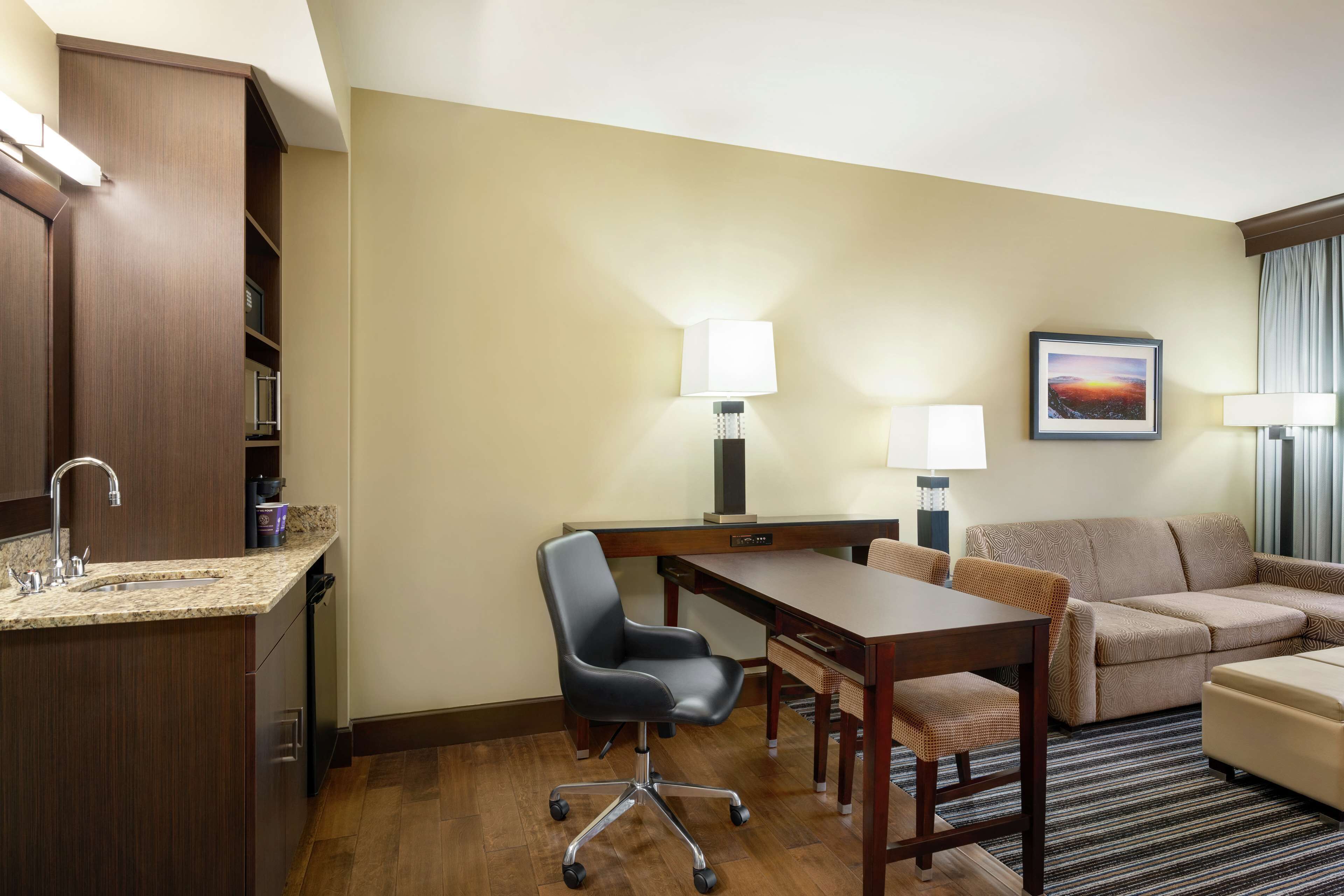 A hotel room with a kitchenette, a desk, a chair, a sofa, and two lamps.