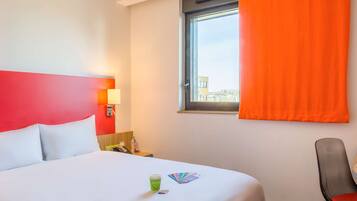 Een hotelkamer met een bed, een raam met oranje gordijnen, een stoel en een nachtkastje met een lamp.