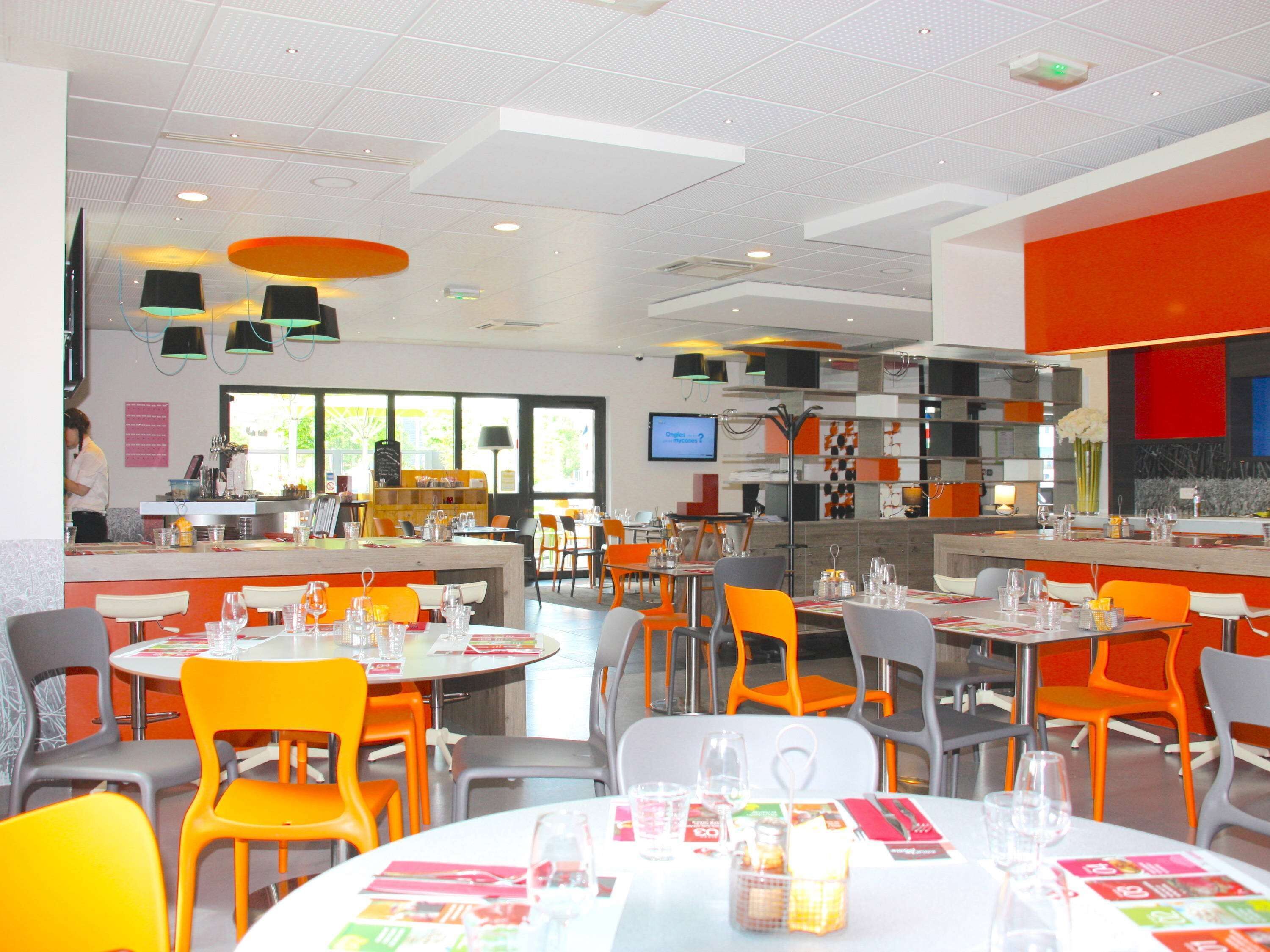 Ein modernes Restaurant mit leuchtend orangefarbenen und grauen Stühlen, weißen Tischen und einer zentralen Bar.