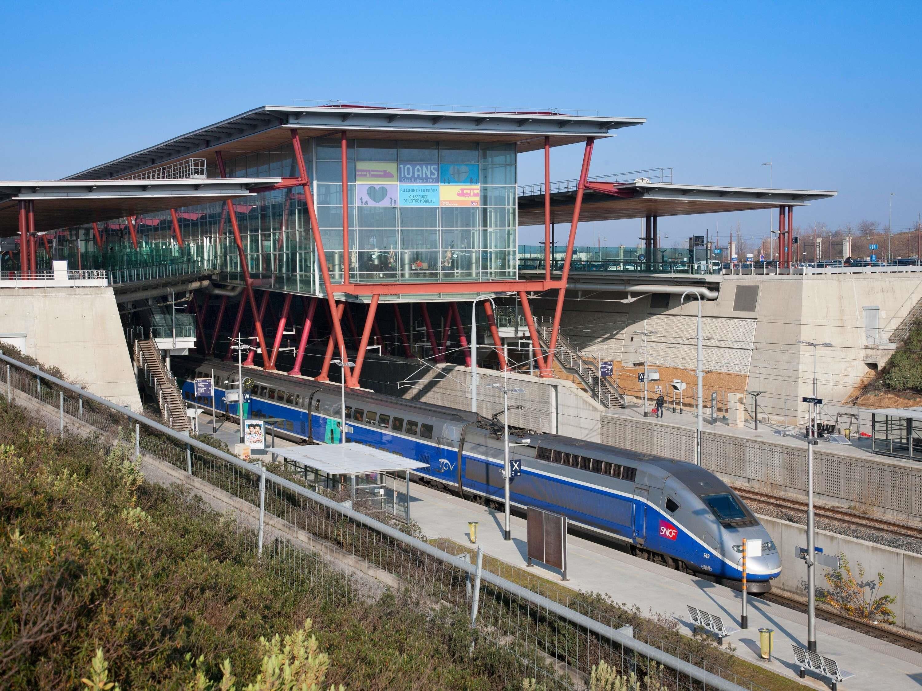 A modern train station with a high-speed train, a glass-walled waiting area, and a red structural framework.