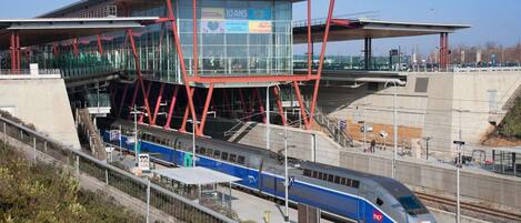 A modern train station with a high-speed train, a glass-walled waiting area, and a red structural framework.