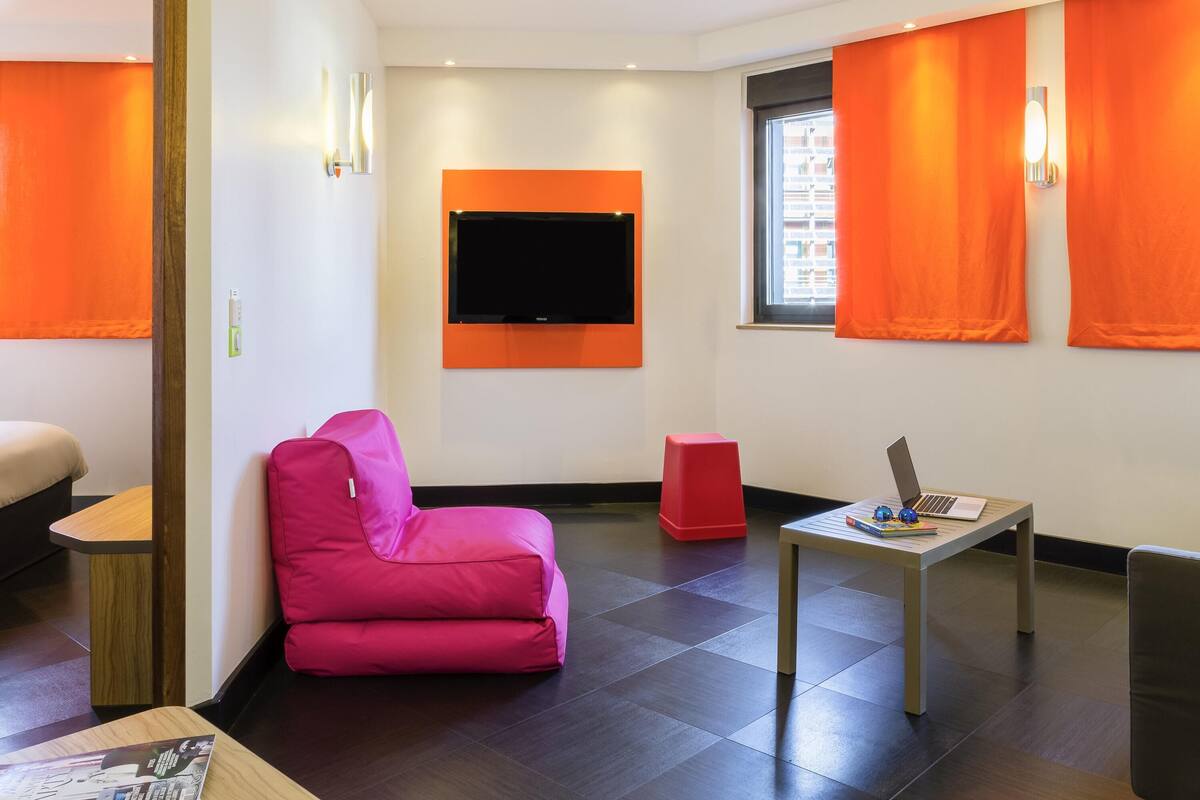 Une chambre d’hôtel moderne avec un téléviseur à écran plat fixé au mur, un canapé rose, un tabouret rouge, une petite table en bois avec un ordinateur portable et des livres, et une fenêtre avec des rideaux orange.