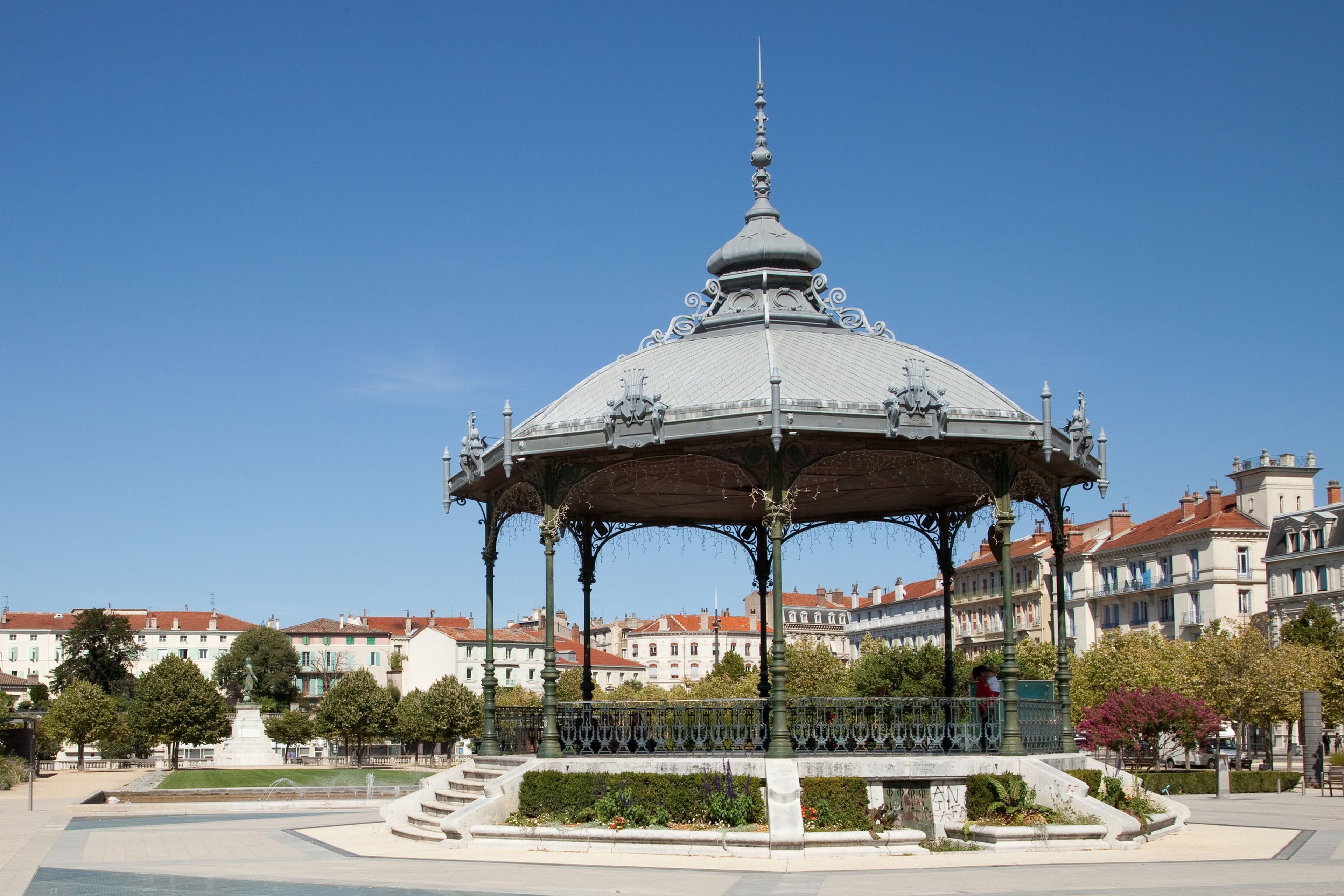 A gazebo with ornate metalwork and a dome roof, surrounded by a landscaped garden and a fountain, with buildings in the background.