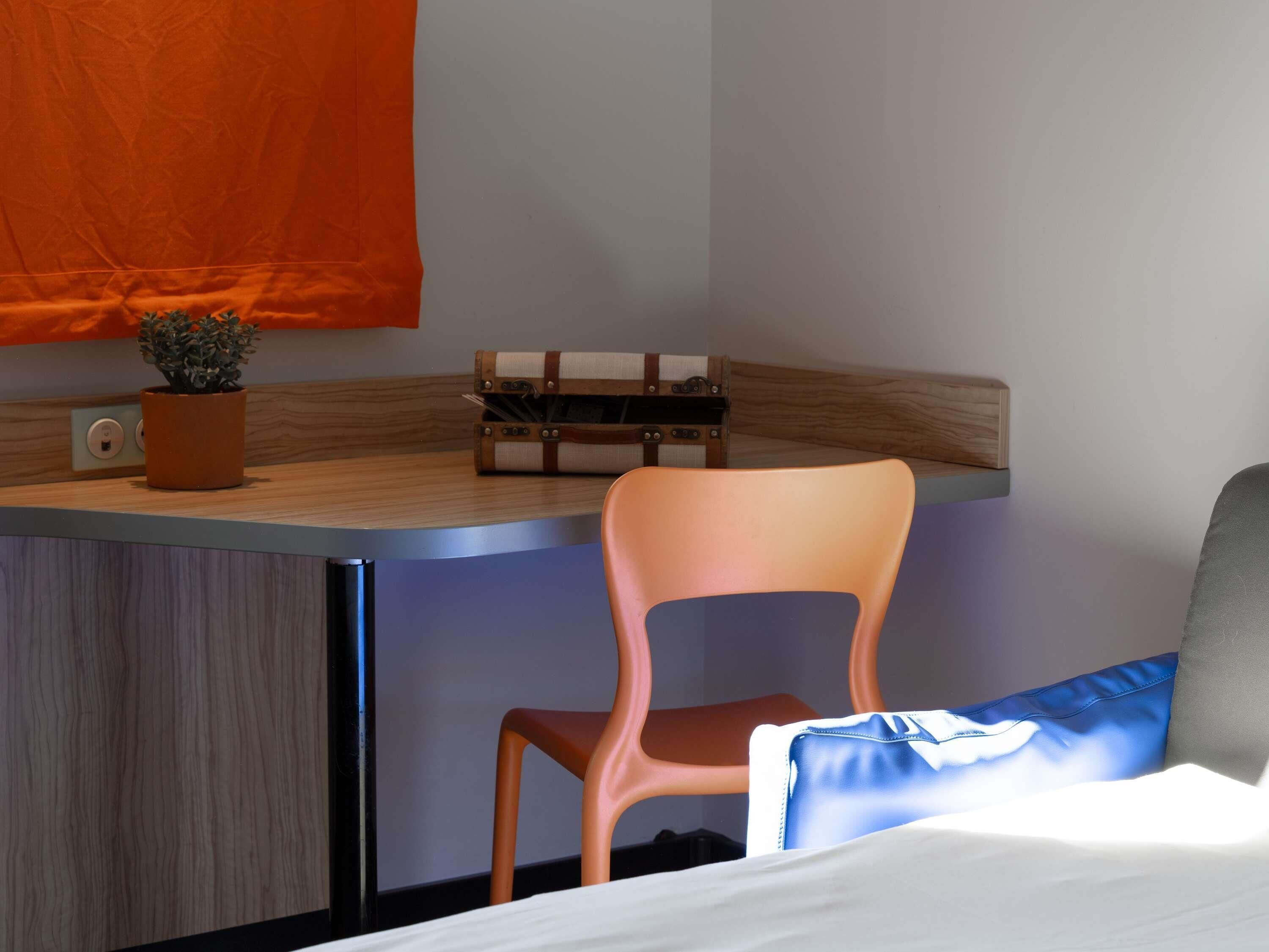 A compact room with a bed, a chair, a small table, a potted plant, and a suitcase.