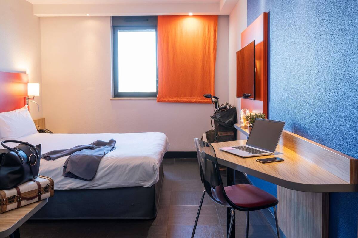 Une chambre d’hôtel avec un lit, un bureau, une chaise et une fenêtre avec des rideaux.
