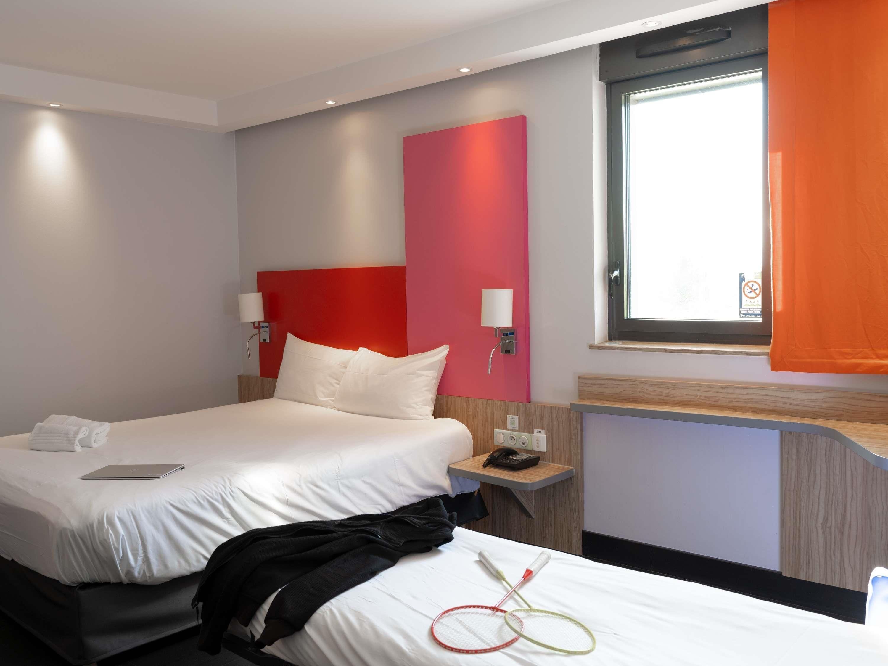 A hotel room with a bed, a bedside table with a phone, a window with curtains, and a wall with a red panel.