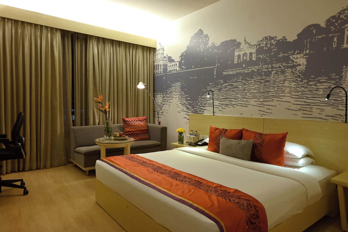 Une chambre d’hôtel avec un grand lit, un canapé, un bureau avec une lampe et une vue sur une rivière, visible à travers une fenêtre avec des rideaux.
