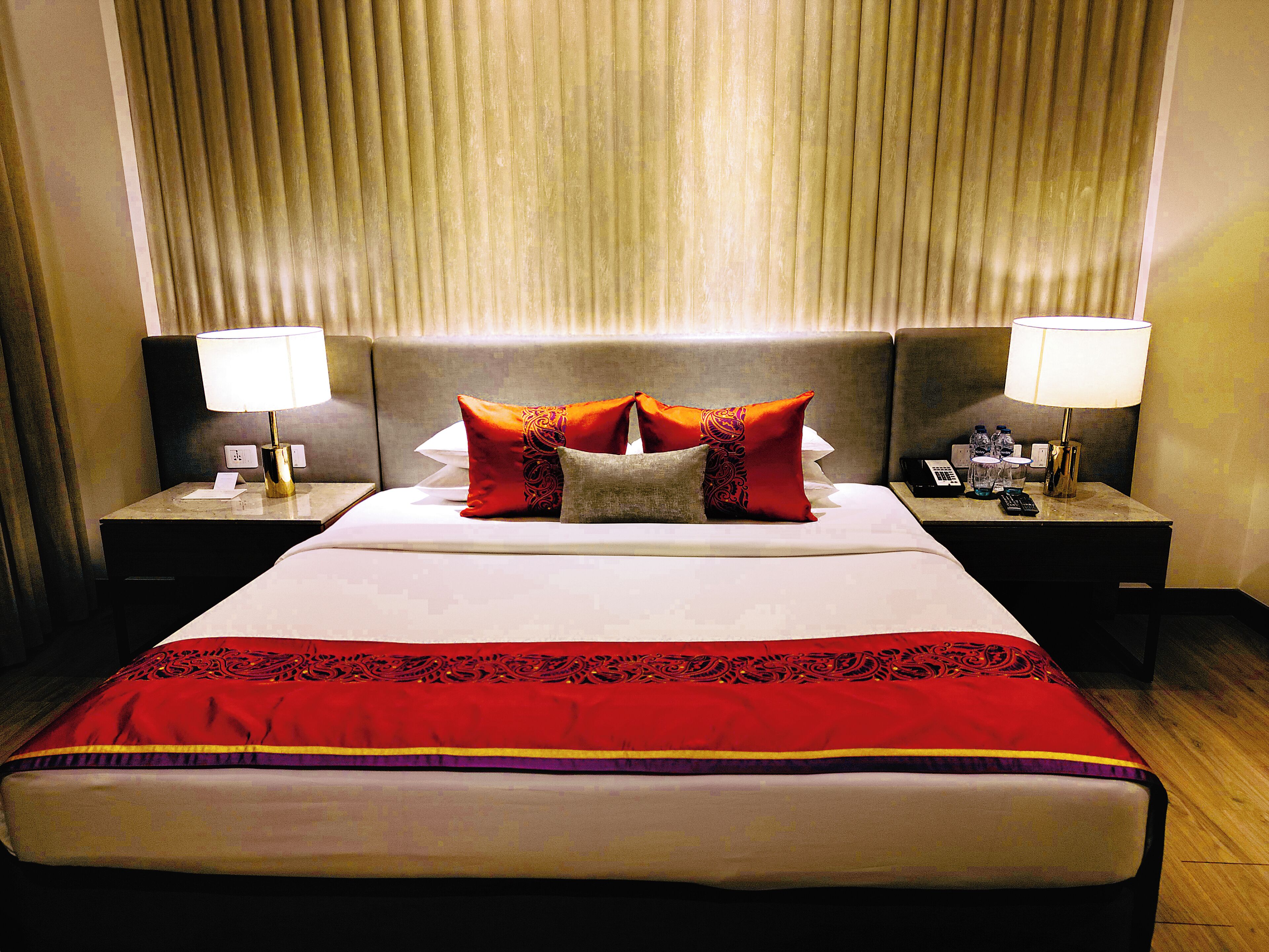 Quarto de hotel com uma cama grande, duas mesas de cabeceira com abajures e uma cabeceira com persianas verticais.