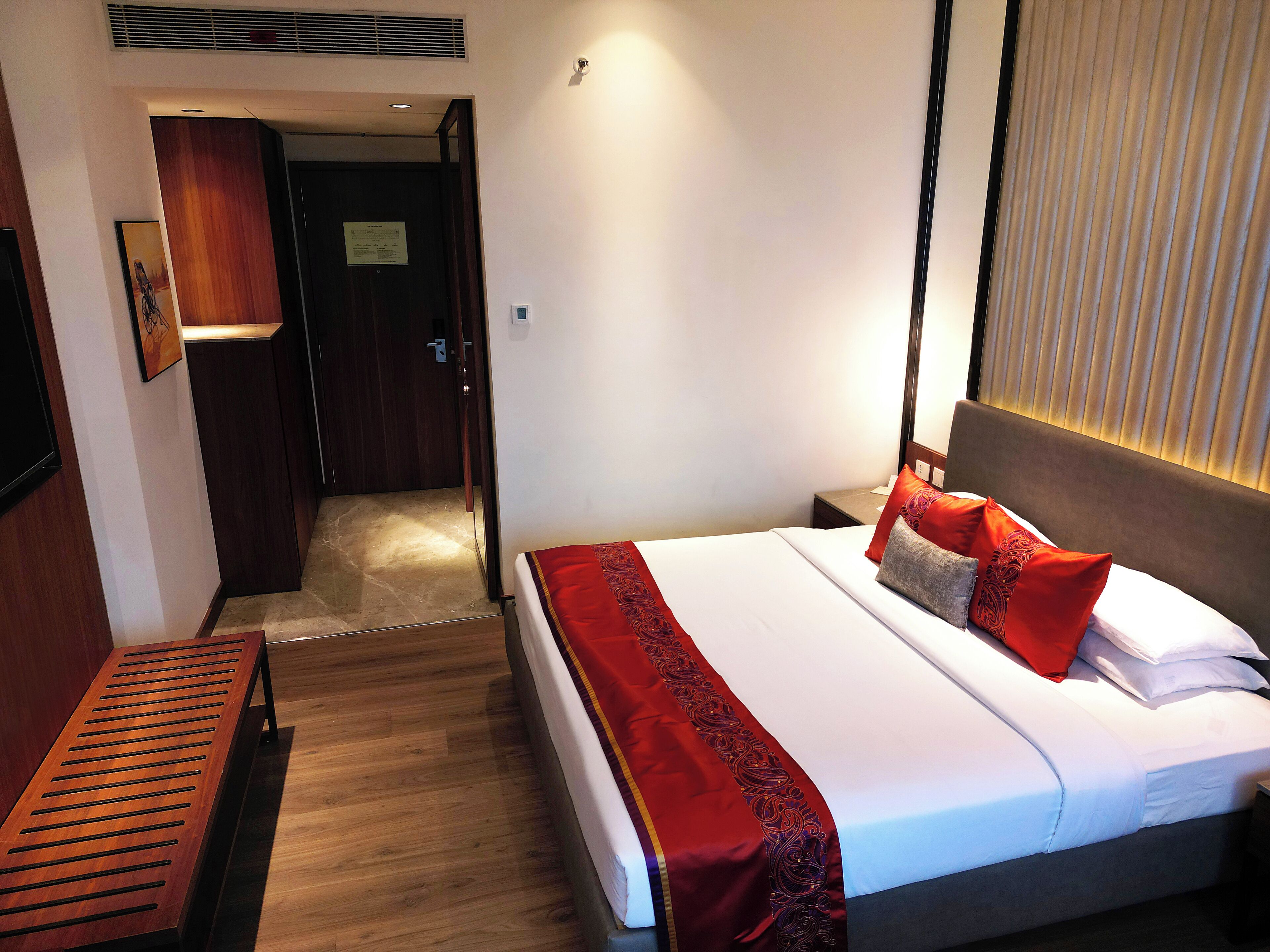 A hotel room with a bed, a wooden bench, a nightstand, and a door leading to another room.
