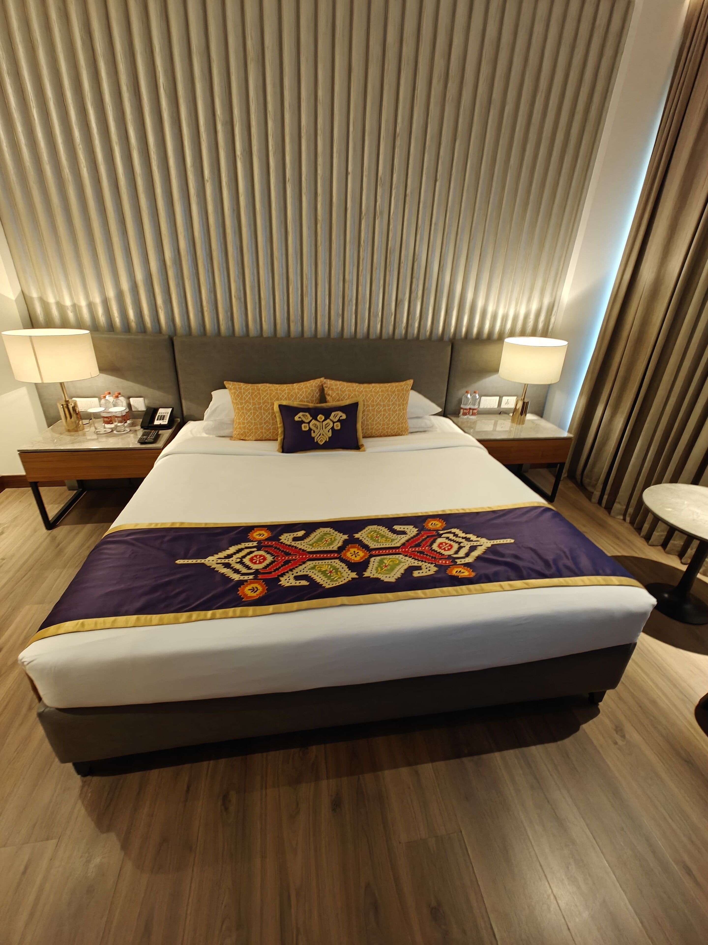A hotel room with a large bed, two bedside tables with lamps, a headboard, and a window with curtains.