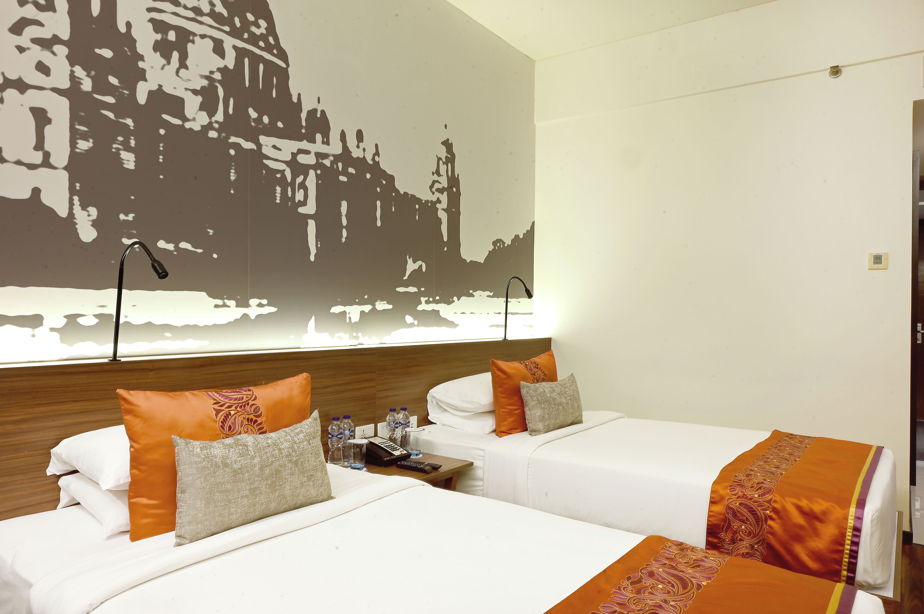 A hotel room with two beds, each with white bedding and orange pillows. There is a nightstand with a remote control, a phone, and bottled water. A large wall mural is visible above the beds.