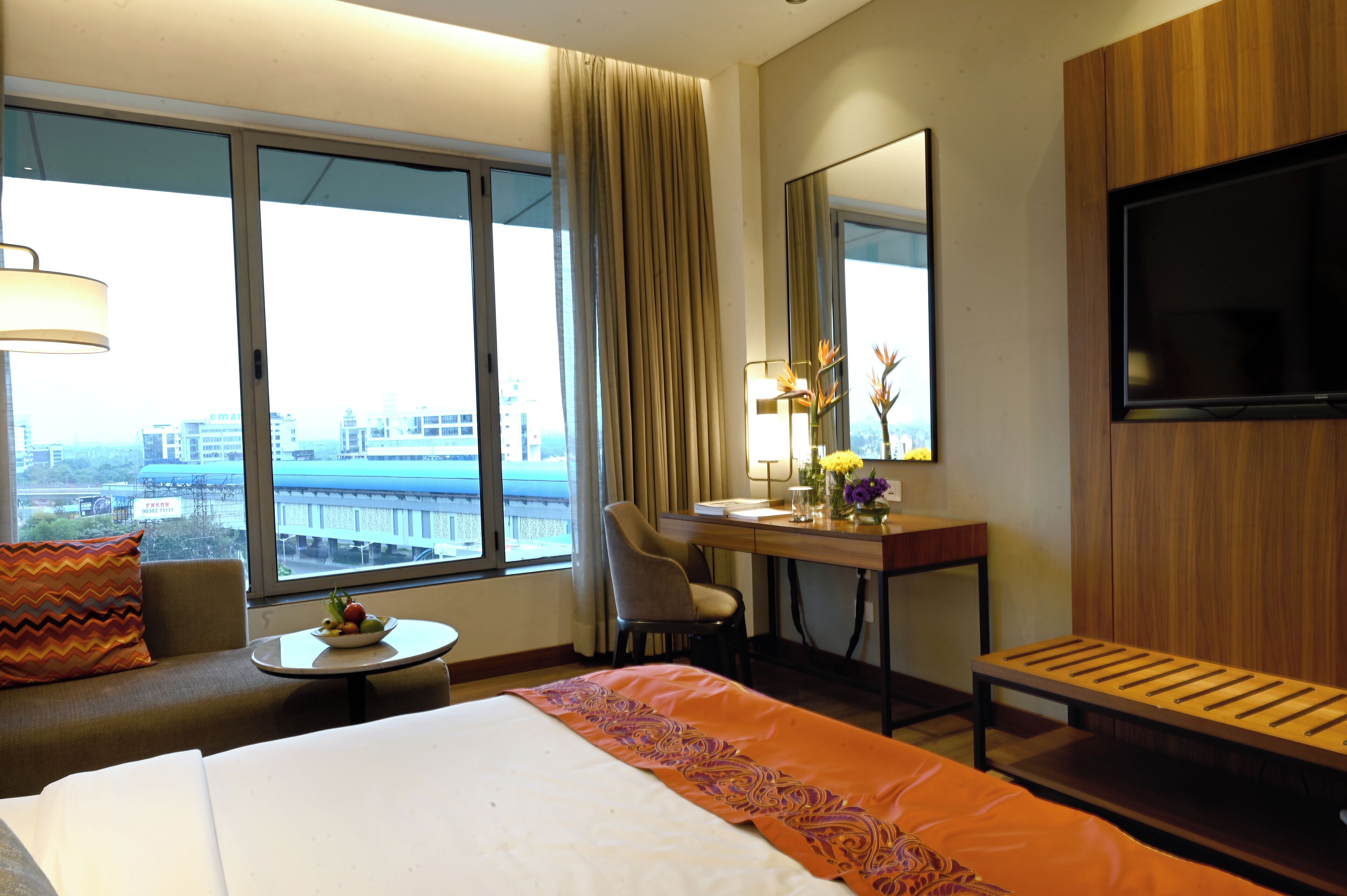 A hotel room with a bed, a desk, a chair, a TV, and a view of a cityscape through large windows.