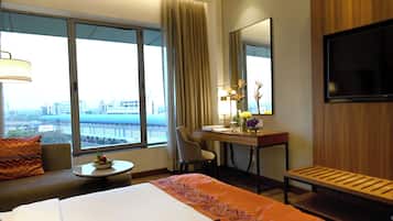 A hotel room with a bed, a desk, a chair, a TV, and a view of a cityscape through large windows.