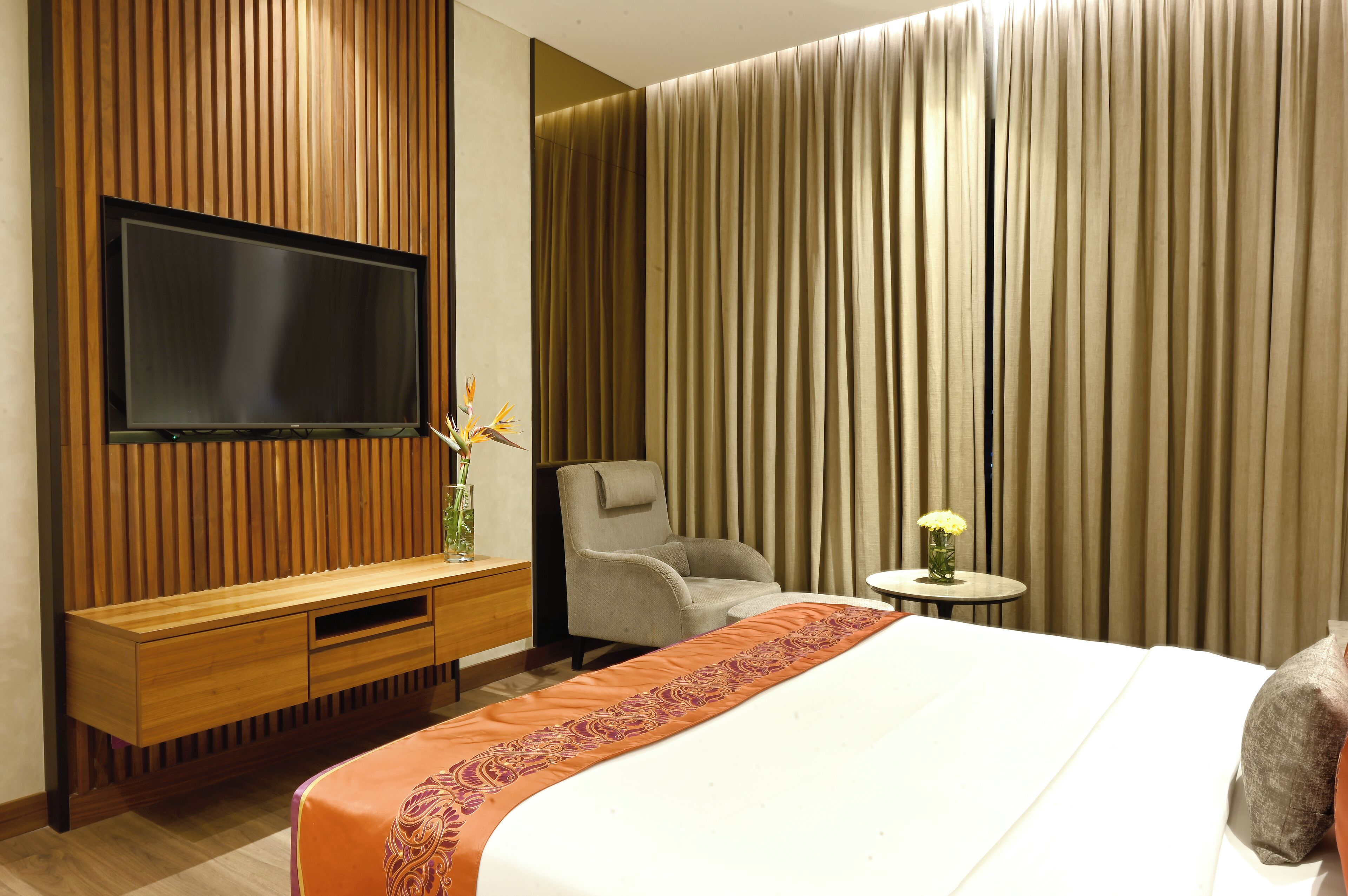Une chambre d’hôtel avec un grand lit, un téléviseur fixé sur un mur recouvert de panneaux en bois, un fauteuil gris et une petite table surmontée d’un vase.