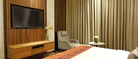 A hotel room with a large bed, a TV mounted on a wooden panel wall, a grey armchair, and a small table with a vase.
