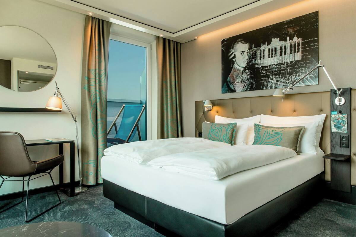Habitación de hotel con una cama grande, un escritorio, una silla, un espejo, una ventana con cortinas y una obra de arte colgada en la pared.