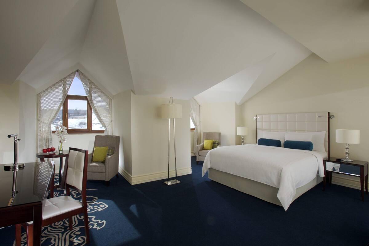 A hotel room with a large bed, a desk with a chair, a window with a view, and a floor lamp.