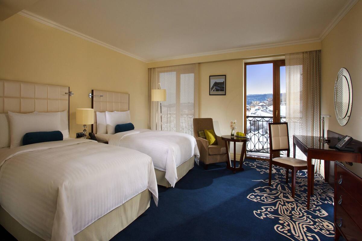 A hotel room with two beds, a desk, a chair, a small table, and a view through a window.