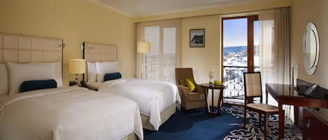 A hotel room with two beds, a desk, a chair, a small table, and a view through a window.