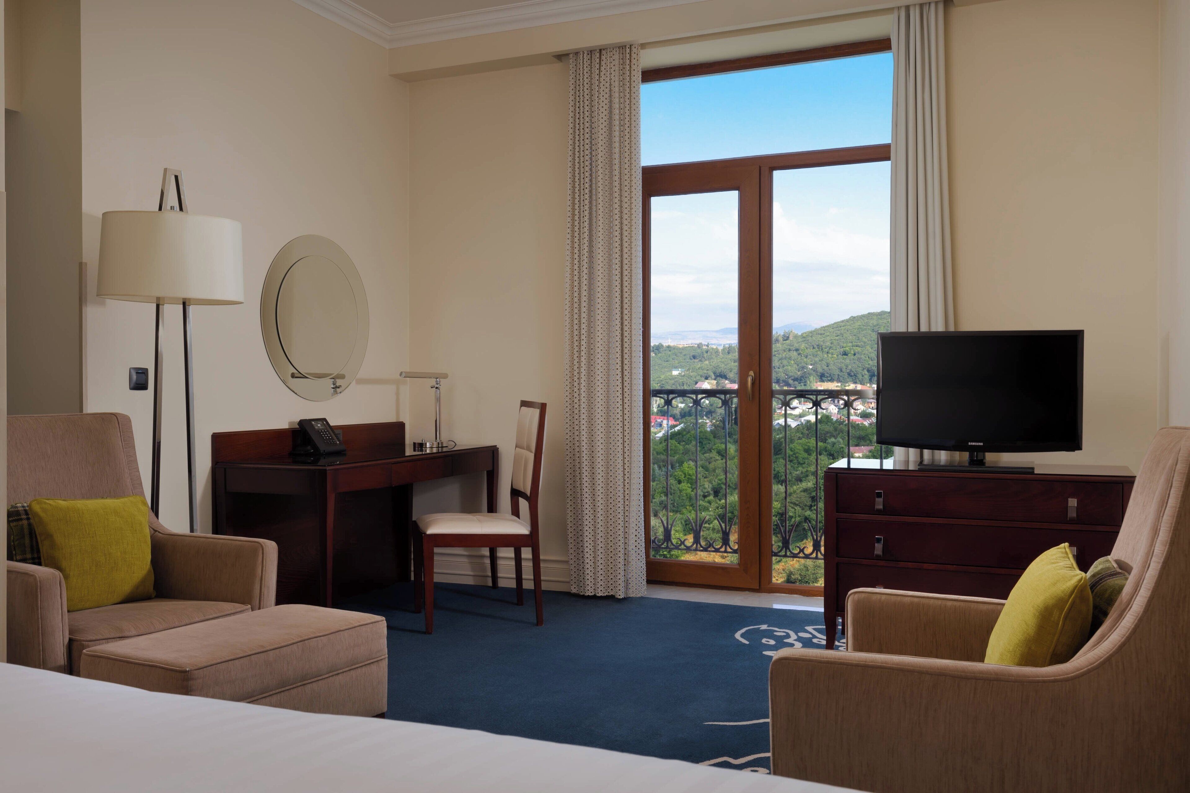 Habitación de hotel con una cama, dos sillones, un escritorio con silla, televisor, cómoda, lámpara, espejo y un ventanal con vistas a zonas verdes y un puente.