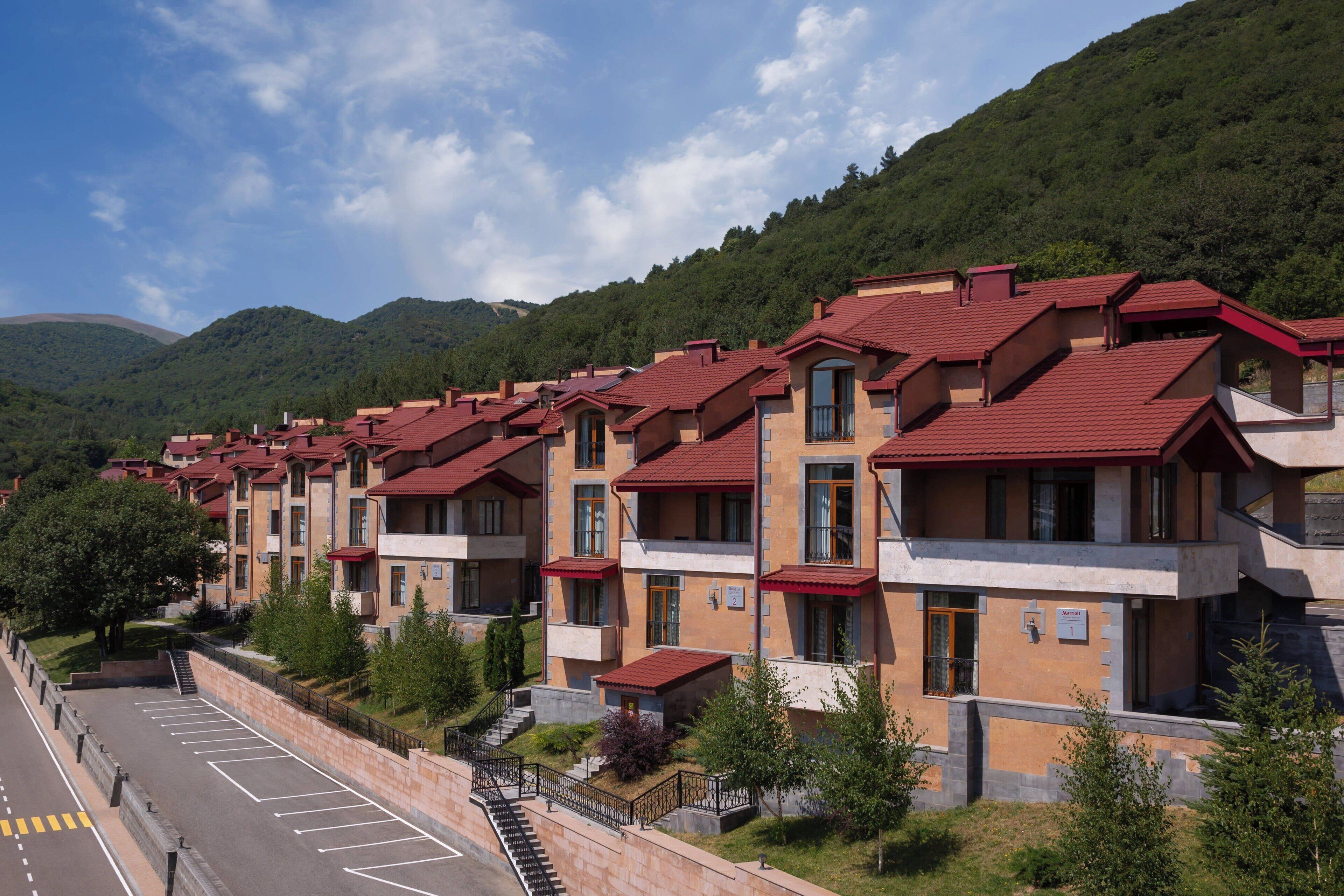 A row of modern apartment buildings with red roofs, set against a backdrop of green hills.