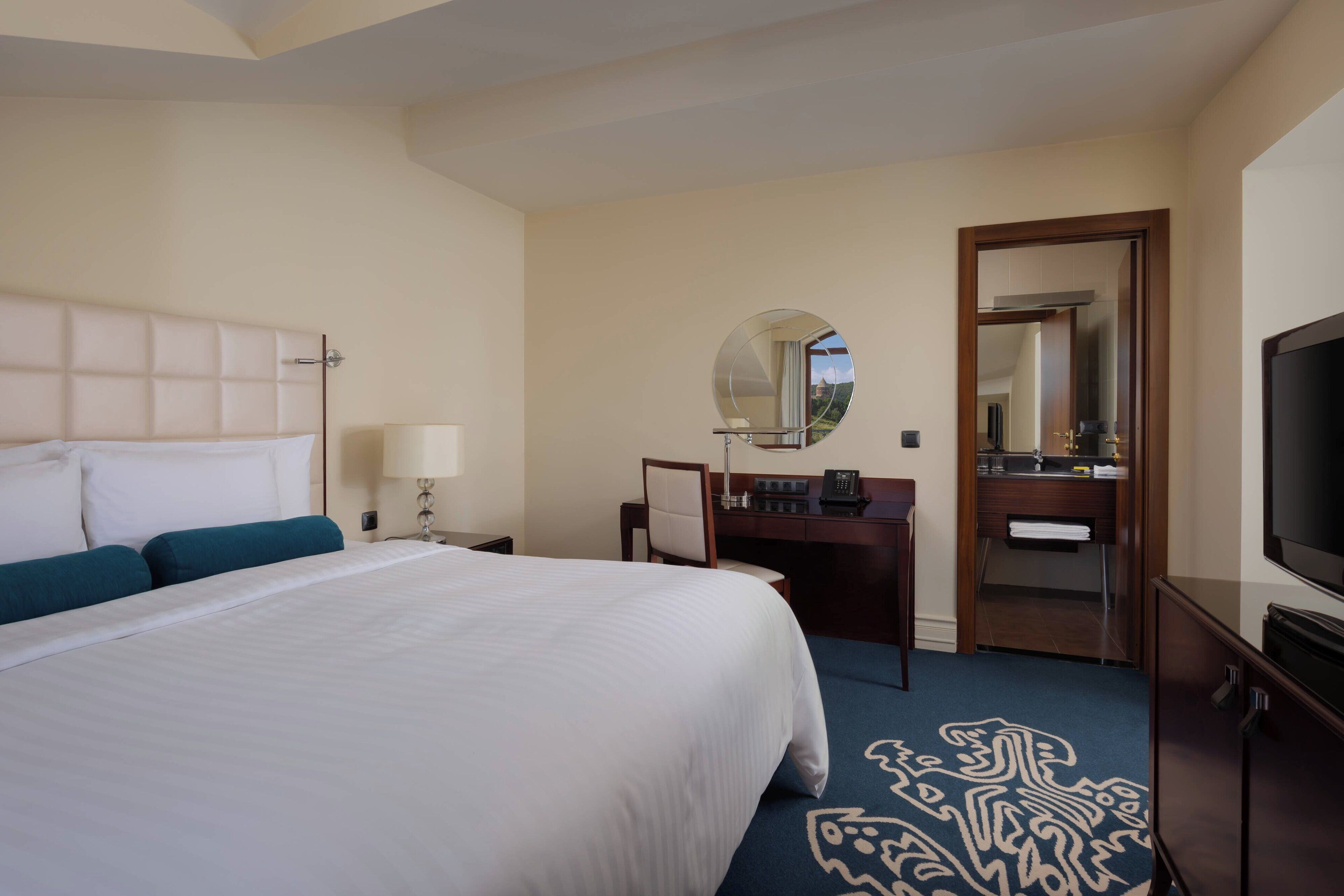 A hotel room with a large bed, a desk with a chair, a mirror, a television, and a bathroom visible through an open door.