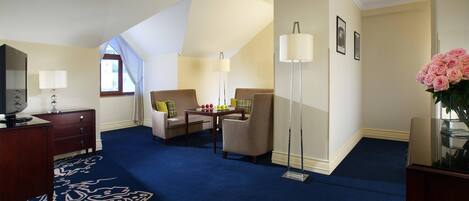 A hotel room with a blue carpet, a desk with a lamp, a sofa, a chair, and a large window.