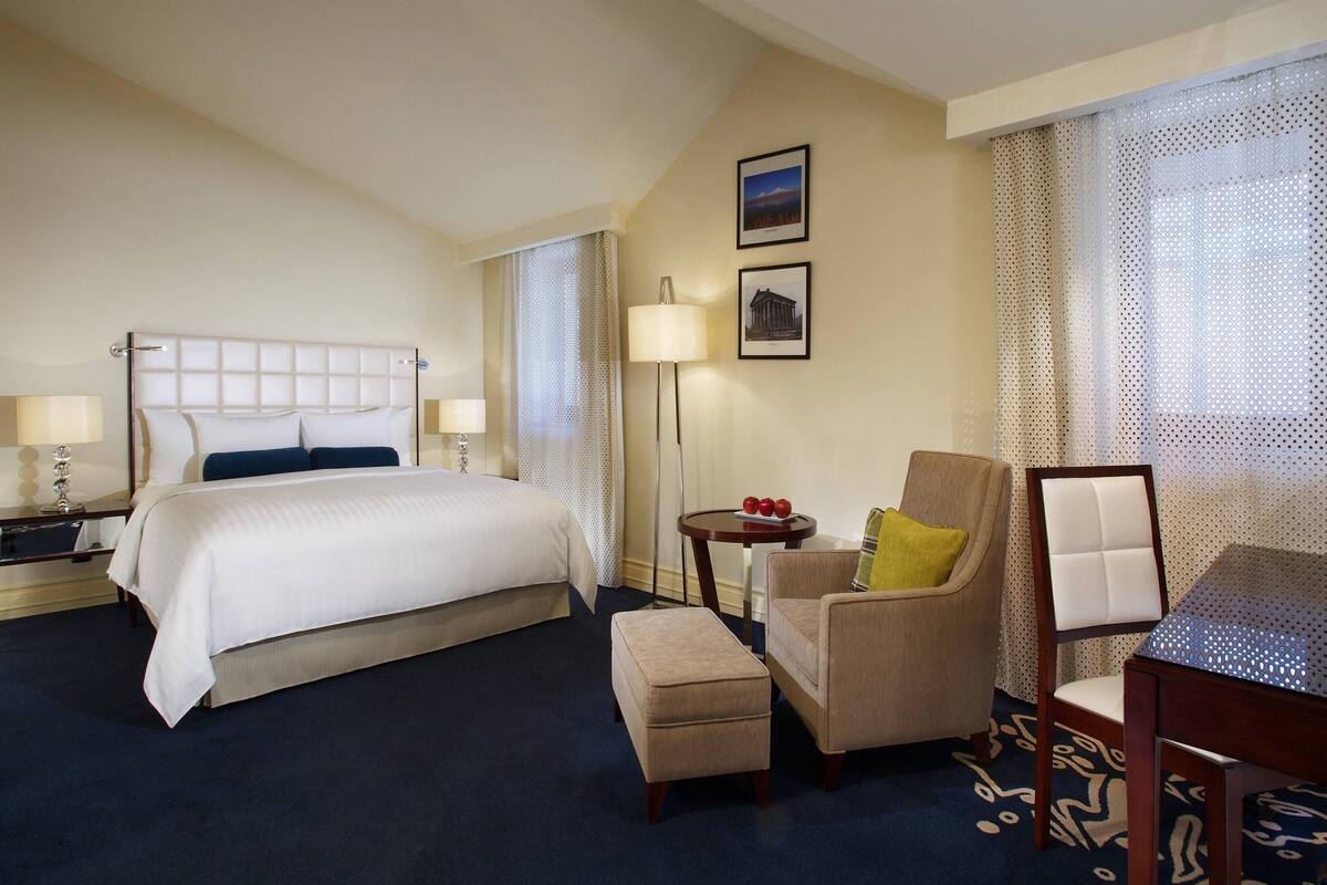 A hotel room with a large bed, a chair, a small table, and a lamp.