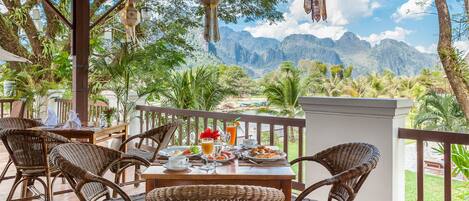 A covered outdoor dining area with wicker furniture and a table set for a meal, overlooking a mountainous landscape.