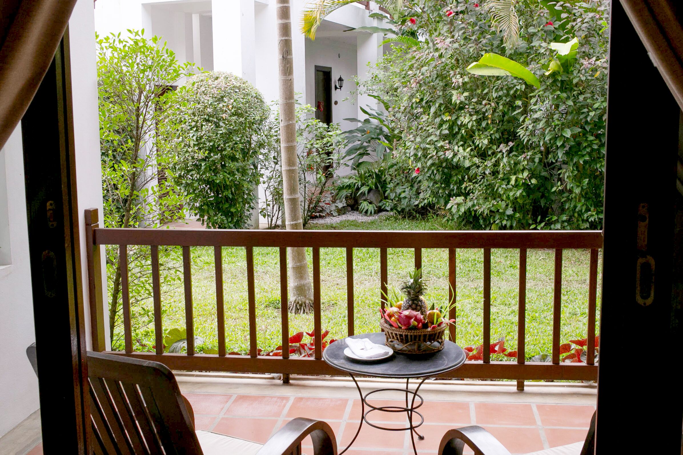 A balcony with a table and chairs, overlooking a garden with various plants and a small pond.
