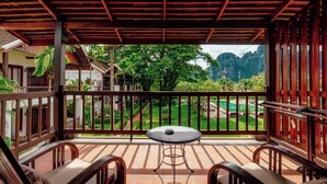 A covered balcony with a table and chairs, overlooking a lush garden and a mountainous landscape.