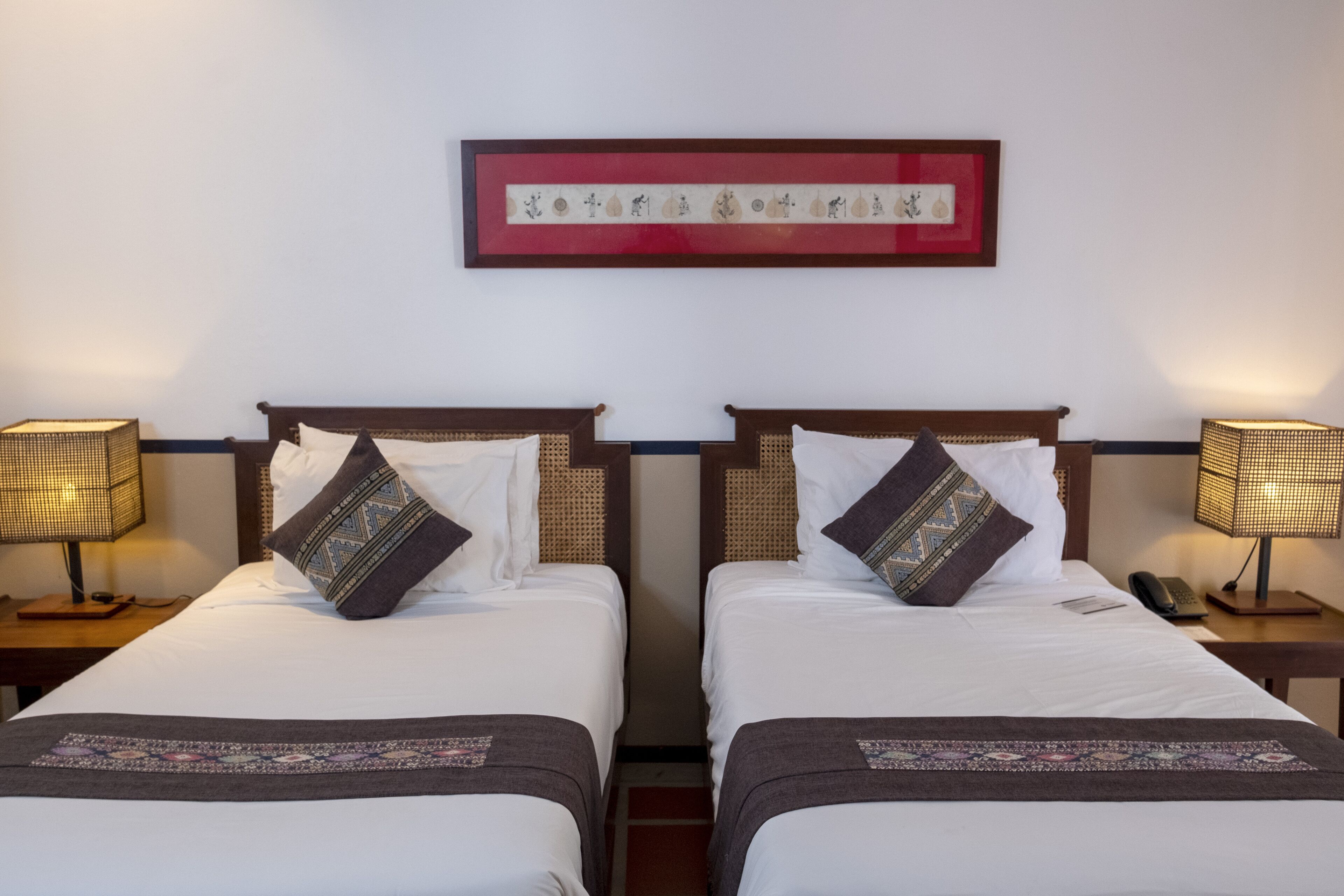 A hotel room with two single beds, each with a patterned throw blanket and pillows. There are bedside tables with lamps, a framed artwork above the beds, and a telephone on one of the tables.