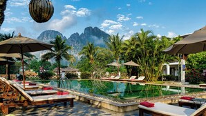 A pool area with lounge chairs and umbrellas, surrounded by palm trees and a mountain backdrop.