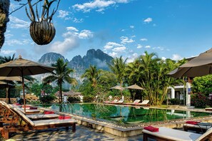 A pool area with lounge chairs and umbrellas, surrounded by palm trees and a mountain backdrop.