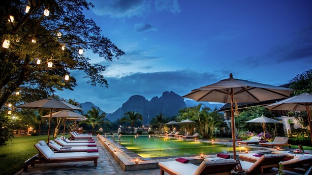Un espace aménagé au bord de la piscine, avec des chaises longues et des parasols, entouré d’arbres et offrant une vue sur la montagne.