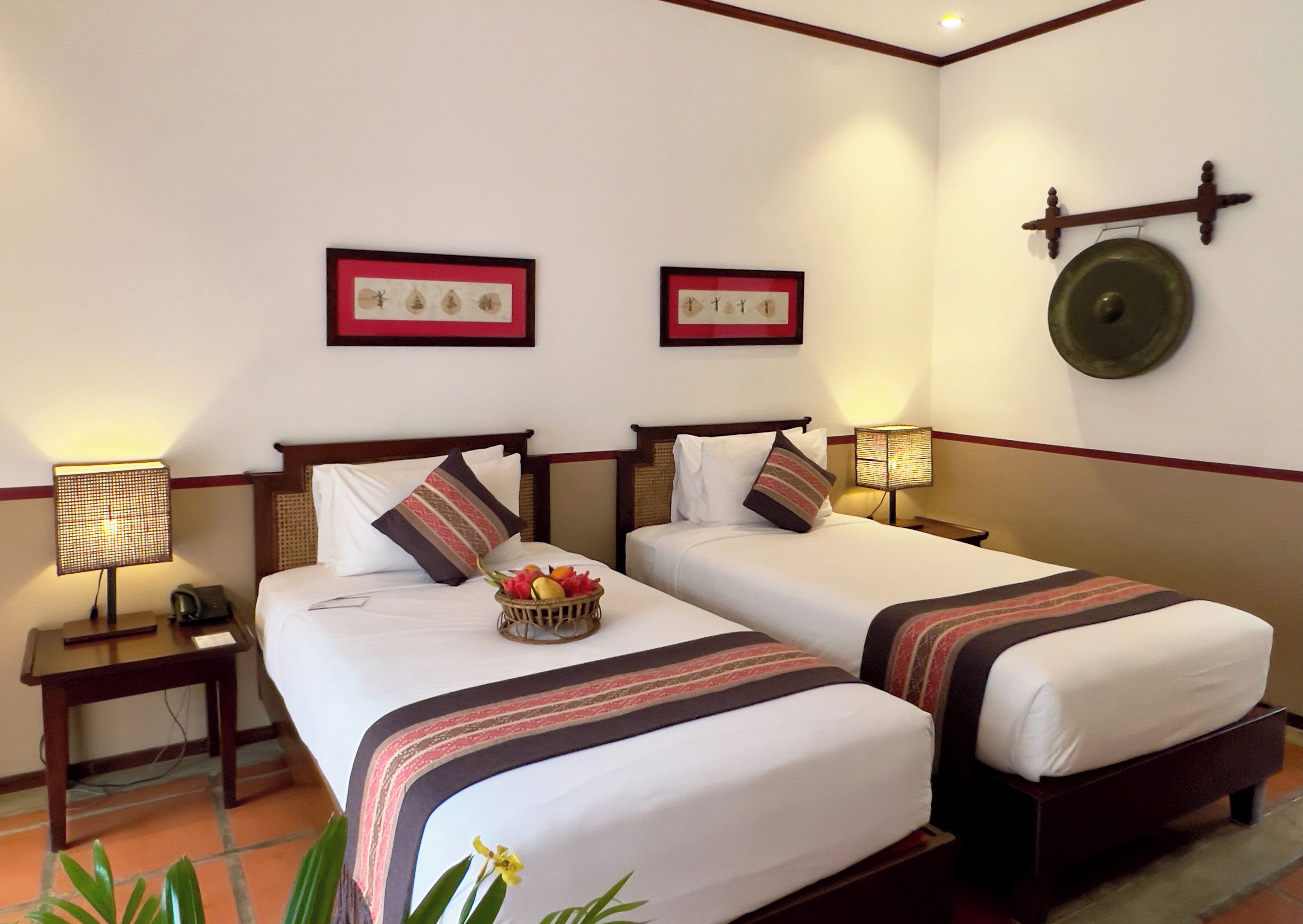 A hotel room with two beds, a bedside table with a lamp, a wall clock, and framed artwork.