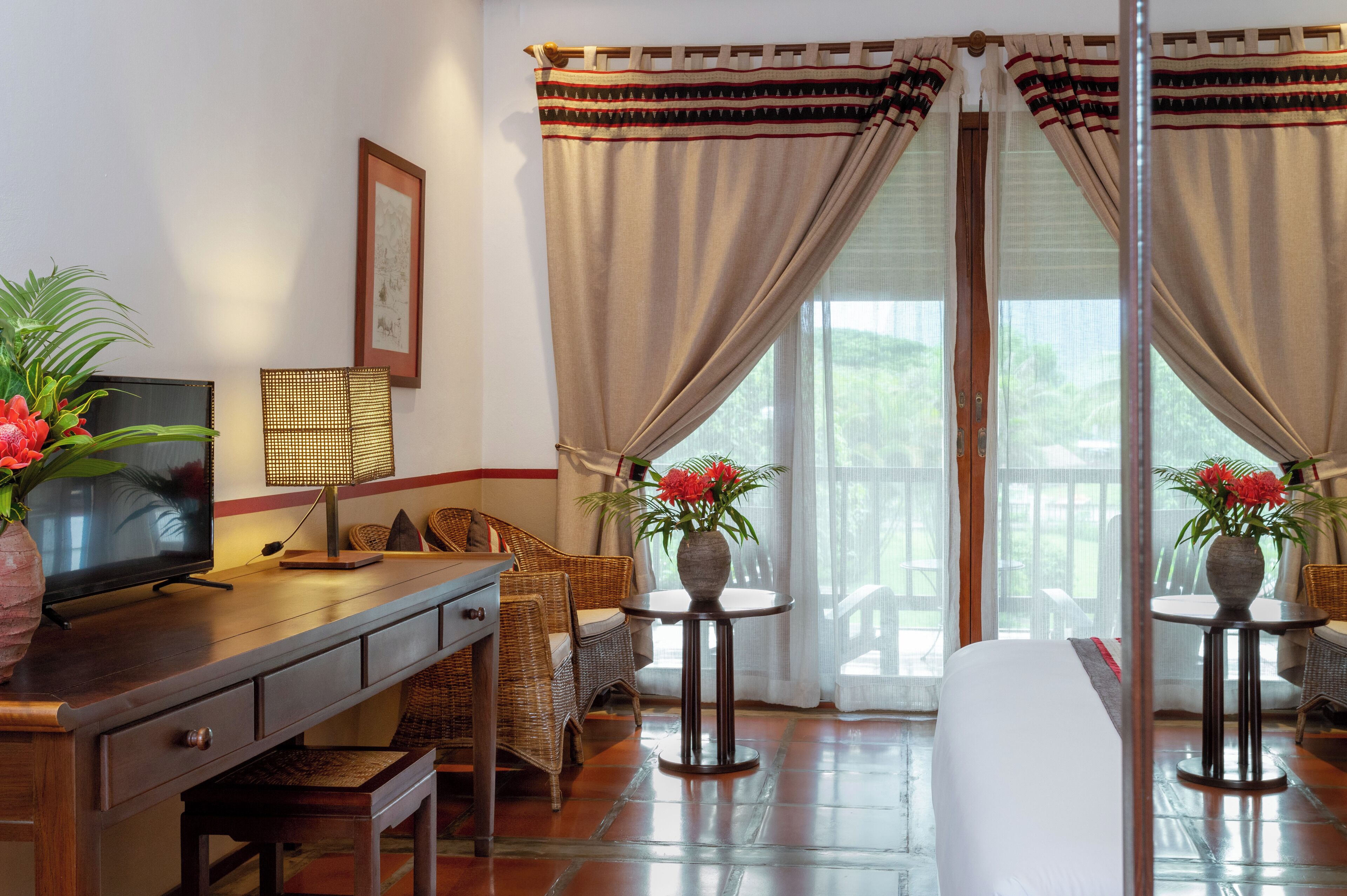 A hotel room with a bed, a desk with a television, a lamp, wicker furniture, and a view through the curtains.