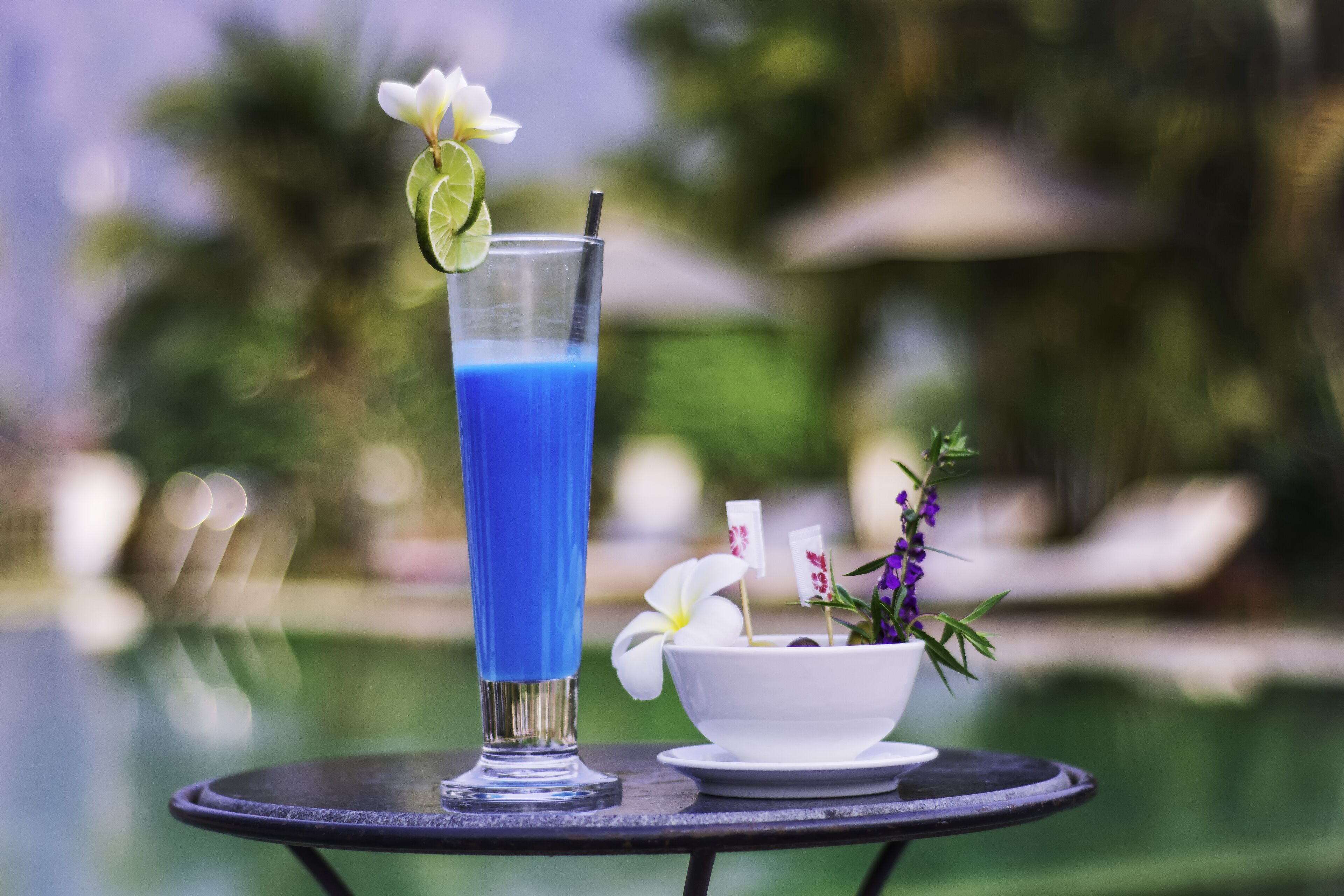Un vaso alto con una bebida azul, un gajo de lima y una pajita, un cuenco blanco pequeño con flores, y una mesa junto a una piscina.