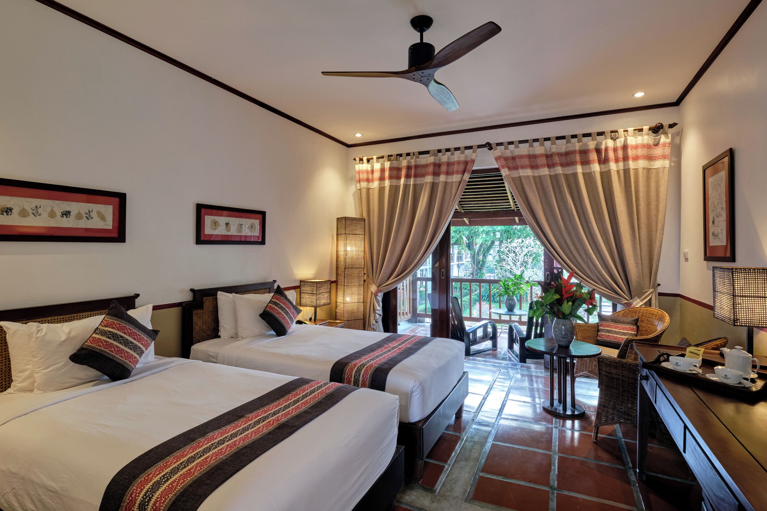 Une chambre d’hôtel avec deux lits, un ventilateur de plafond, des rideaux, une petite table avec un vase et une vue sur l’extérieur.