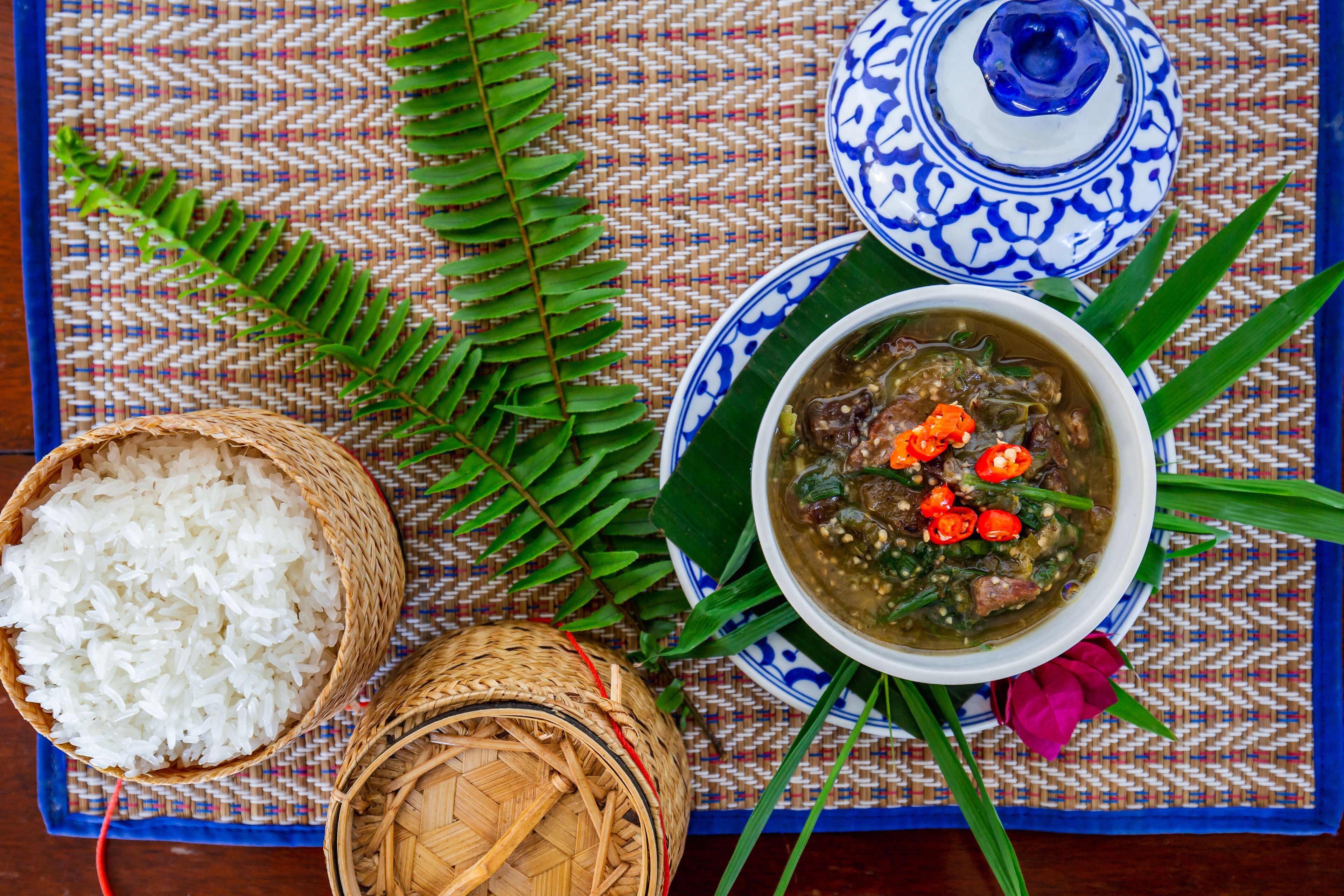 Eine gewebte Matte mit einem gewebten Korb voller weißem Reis, einer Schüssel Suppe mit roten Chilischoten und einem blau-weißen Keramiktopf.