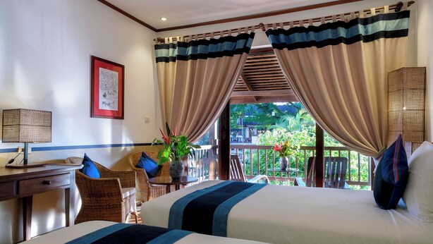 A hotel room with two beds, a desk, wicker chairs, a potted plant, and a balcony with a view of greenery.