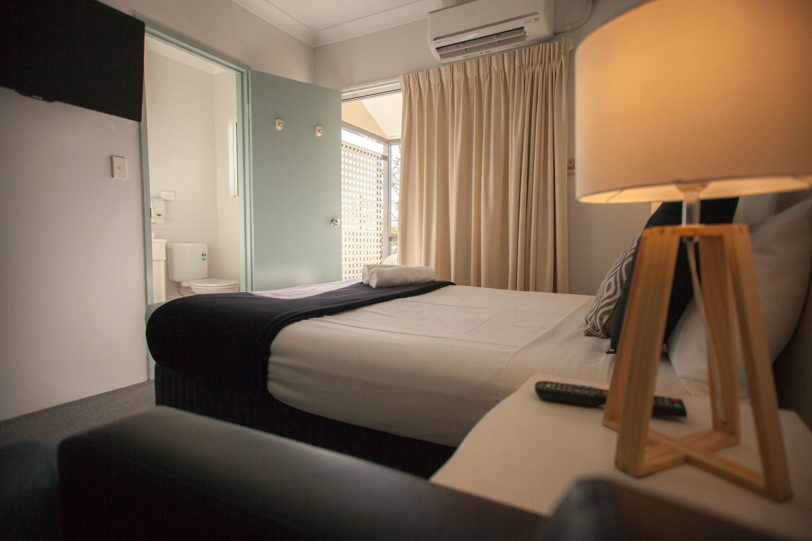 Habitación de hotel con cama, mesita de noche, lámpara y vista al exterior a través de una ventana con cortinas.