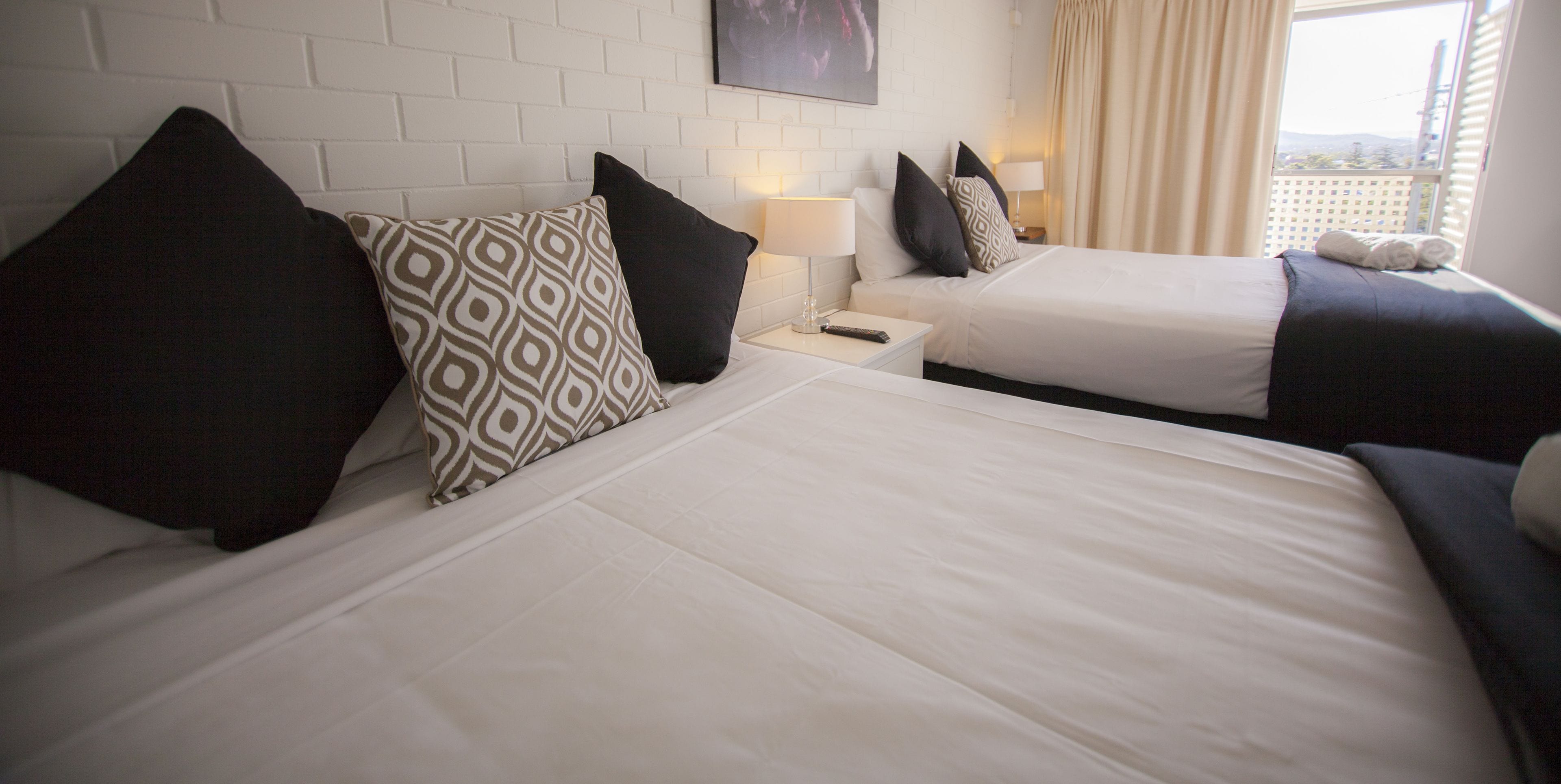 A hotel room with two beds, white bedding, patterned pillows, bedside lamps, and a window with curtains.