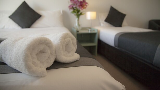 A hotel room with two beds, each with a pillow and a rolled white towel. There is a nightstand with a lamp and a vase of flowers between the beds. - Chermside Court Motel (Chermside)