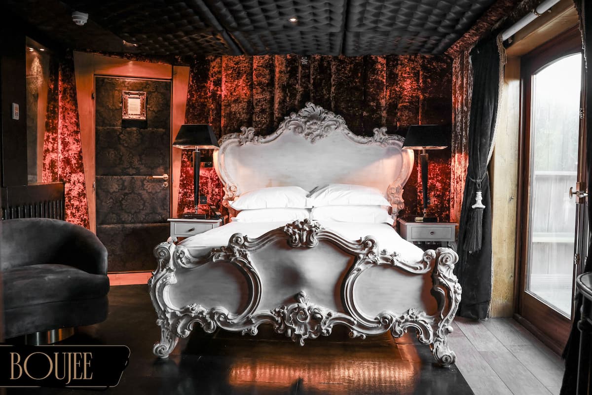 A luxurious bedroom with an ornate bed, two bedside tables, a chair, and a large window.