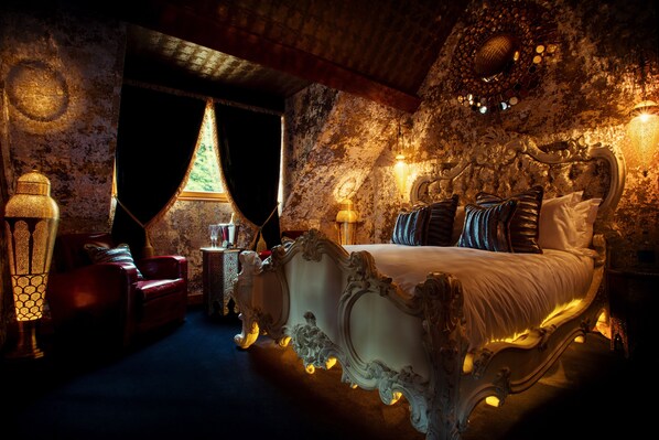 A luxurious bedroom with a large ornate bed, a red leather armchair, and a chandelier.